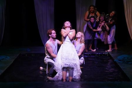 Seven students on stage with water at the center. One student falls back on two with arms dramatically crossed over her chest. A crowd in the back clusters together